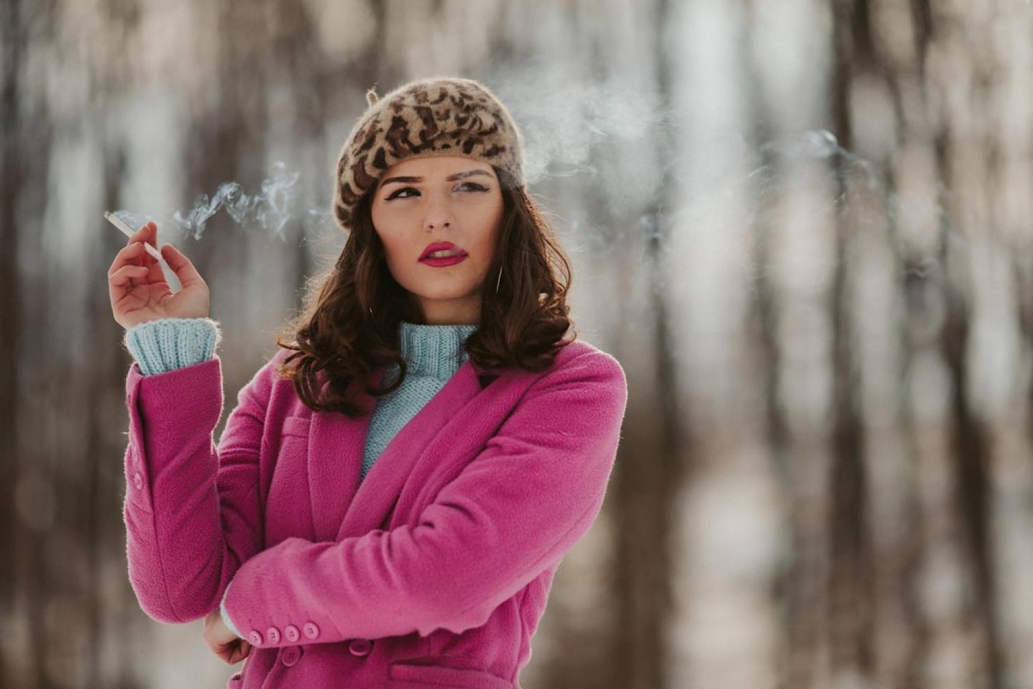 Woman in pink coat smoking a cigarette in a wintery forest, evoking mid-century style